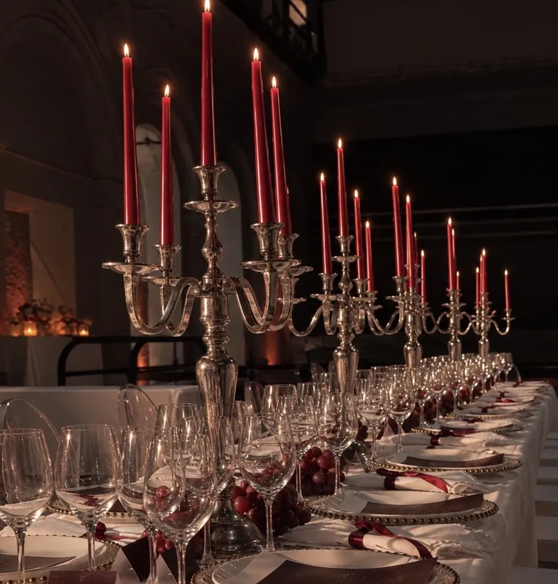 Mesa larga con candelabros de plata, velas rojas encendidas, copas y lazos vino